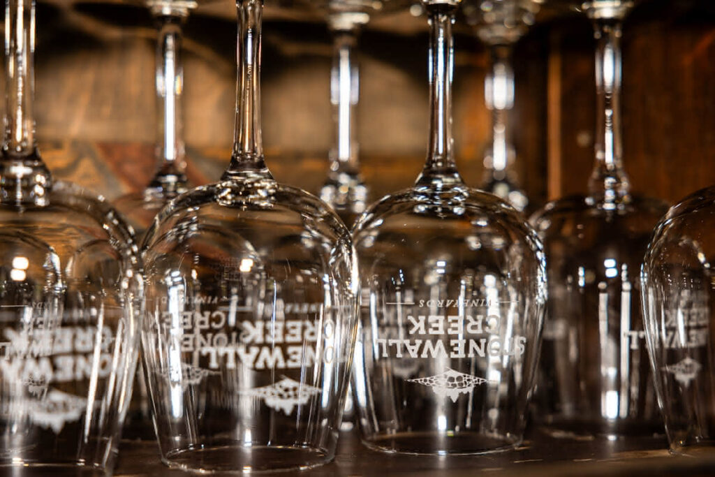 A closeup of several wine glasses upside-down on a dark counter. The glasses have the words “Stonewall Creek Vineyards” printed on them with a small image of a field and trees.