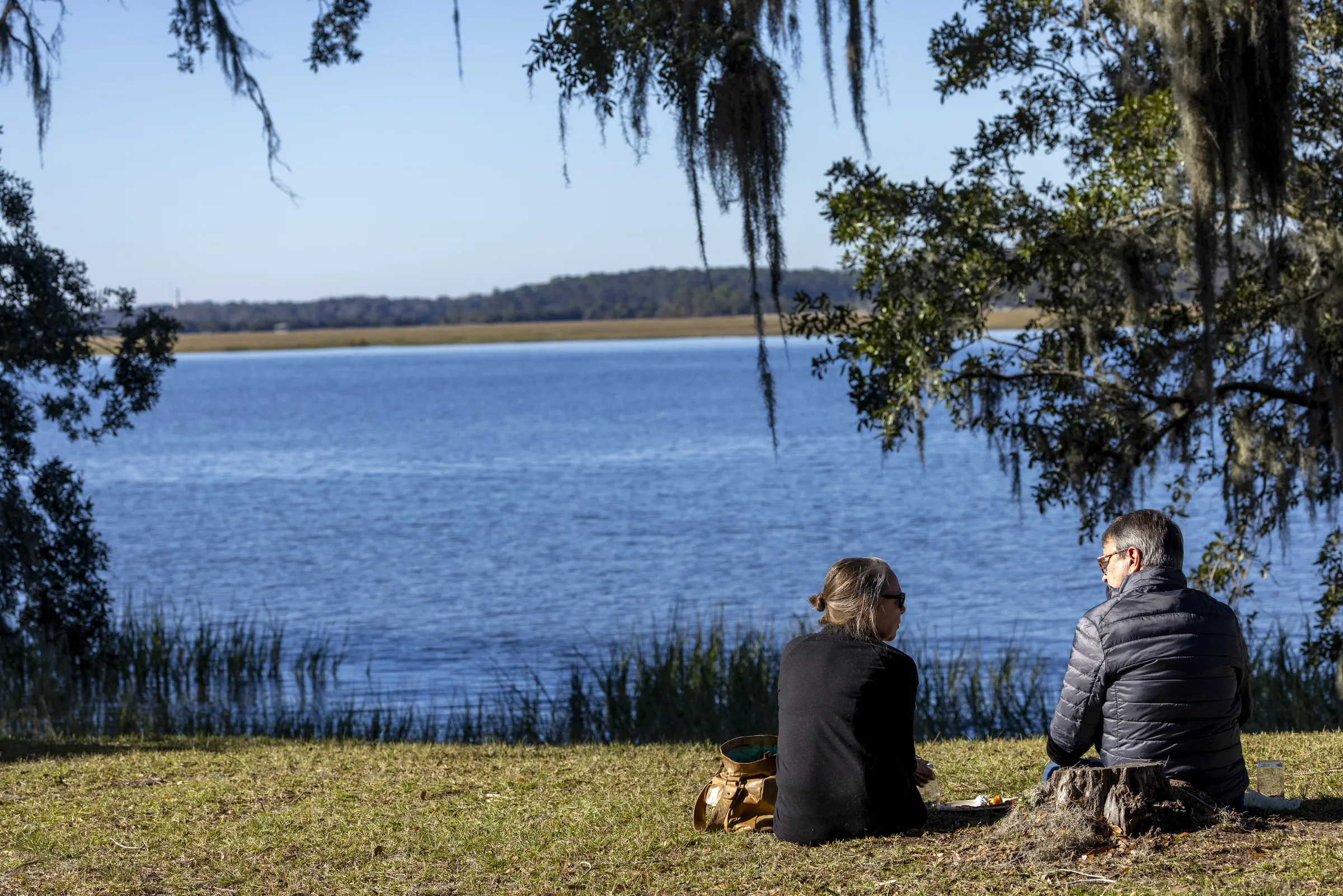 A man and woman sit on the grass and talk to each other with their backs to the camera. In front of them is a large, sparkling blue lake.