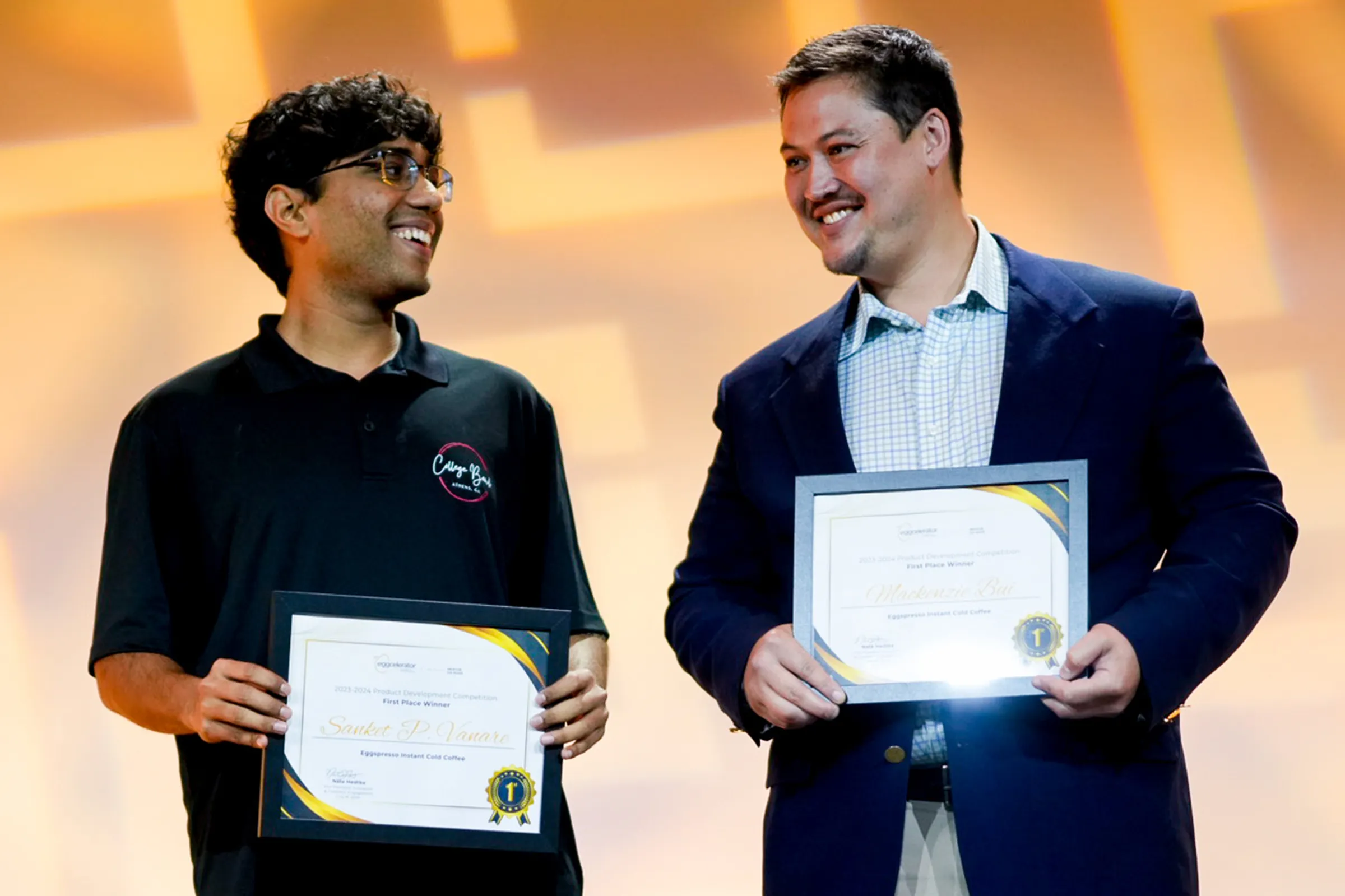 Two young men hold award certificates after winning a product development competition. They are smiling at each other in front of a bright orange background.