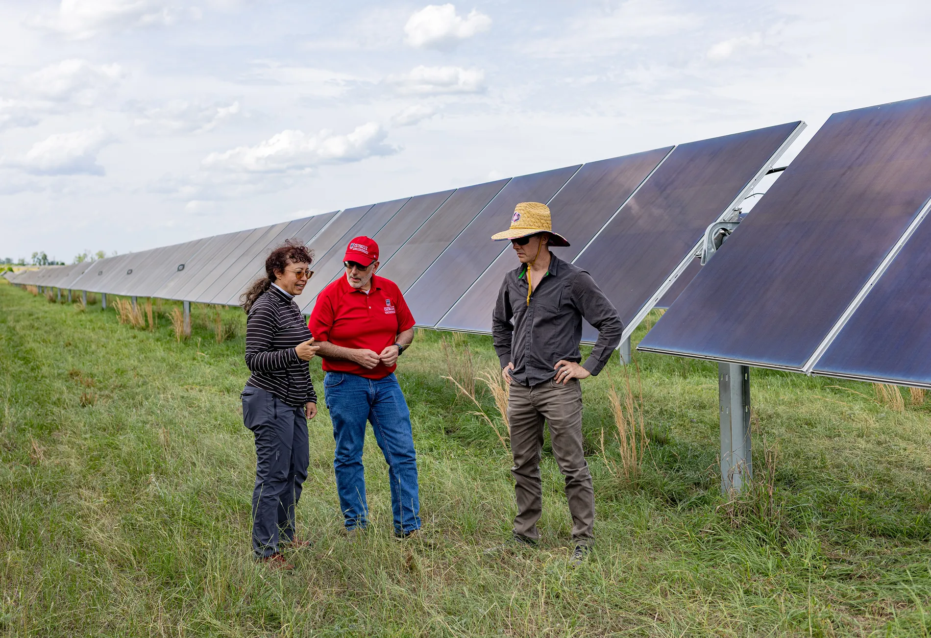 Three people in casual clothing stand in grassy fields next to a long row of solar panels.