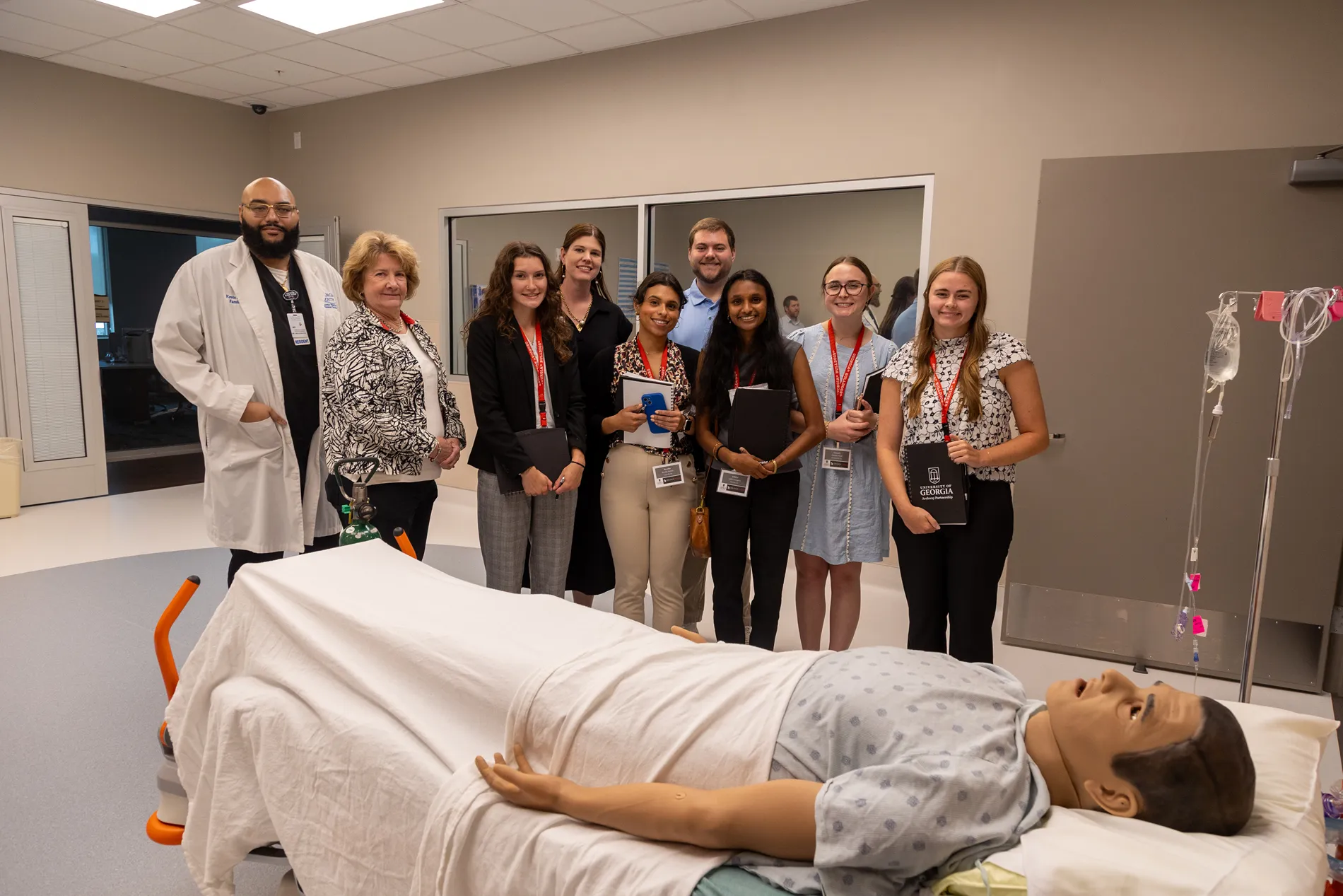 A group of medical professionals and students pose for a photo while standing behind a medical dummy in a bed.
