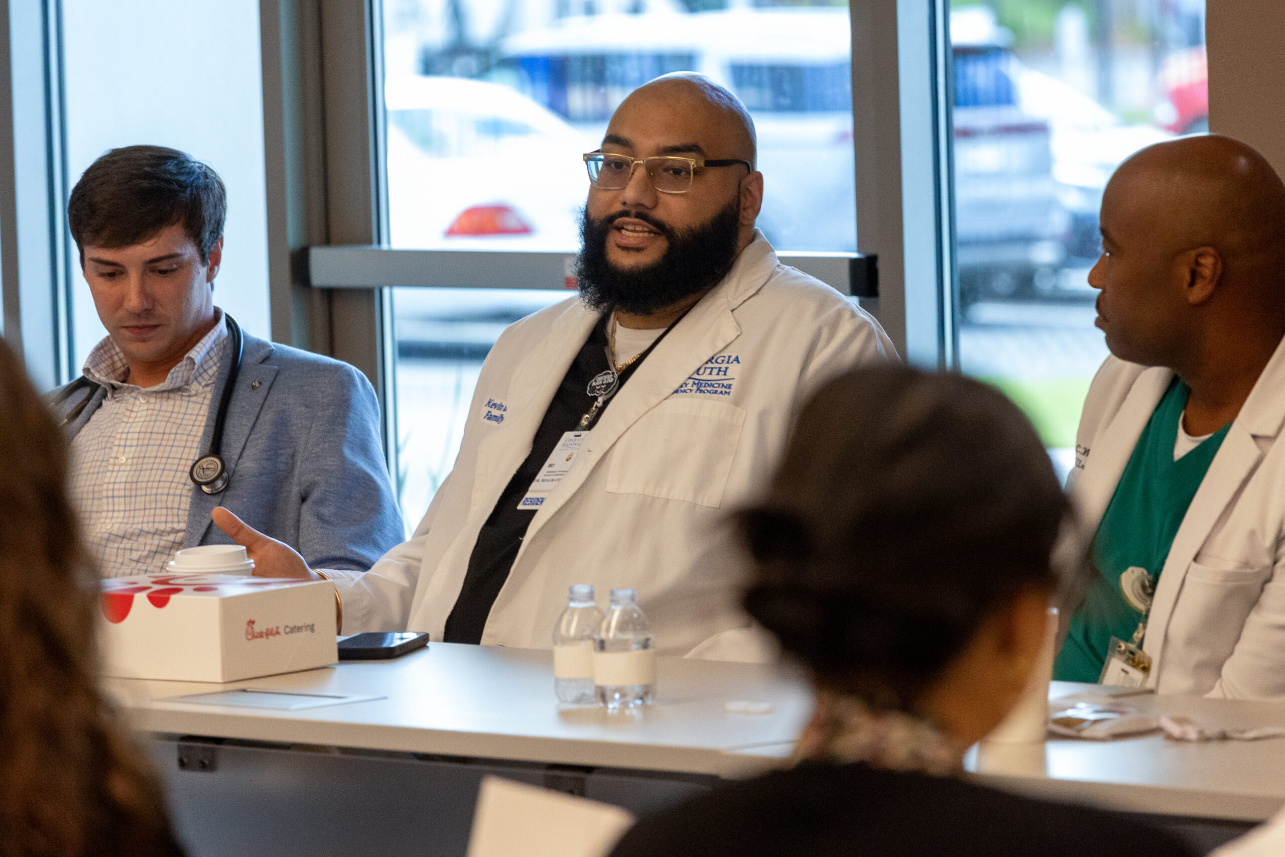 A man in a white medical coat sits at a table and speaks to an audience.