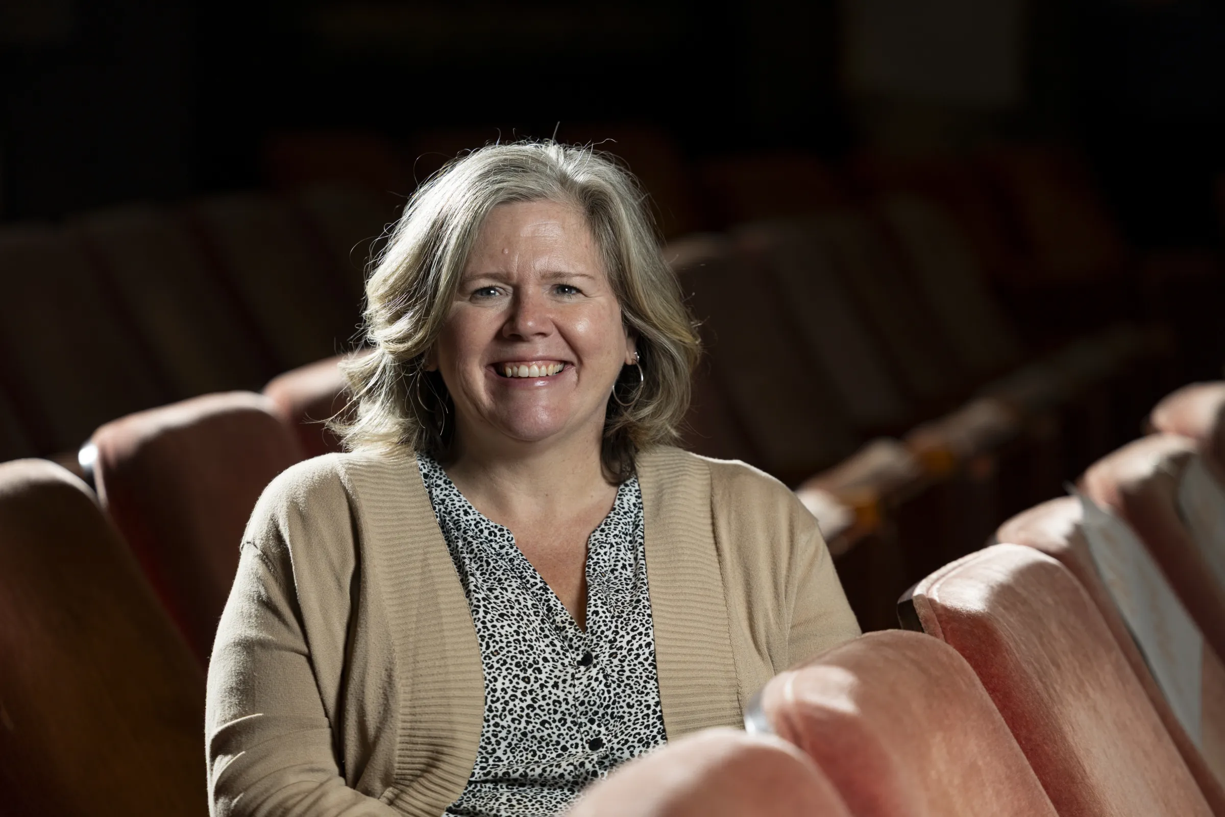 A woman in a tan cardigan smiles while seated in a row in a dramatically lit theatre.
