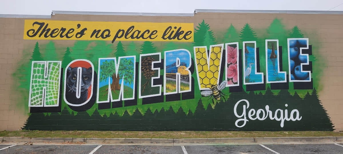 A colorful outdoor mural painted on a brick wall reads "There's no place like HOMERVILLE Georgia," with each block letter displaying a local scene or symbol.